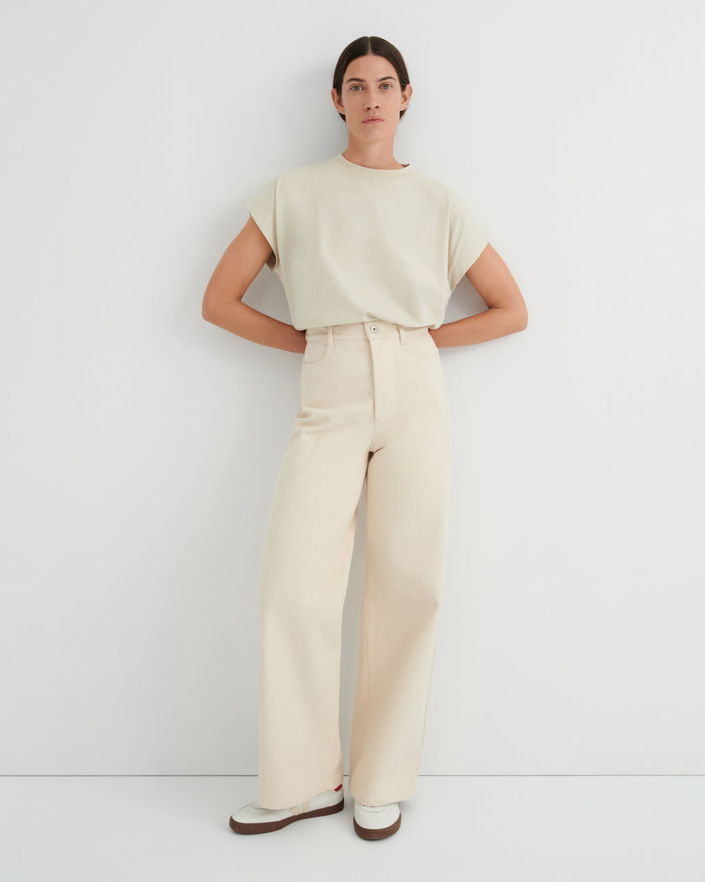 A woman modeling a light cream short-sleeve crewneck top and matching high-waisted wide-leg trousers, standing against a plain white studio wall.
