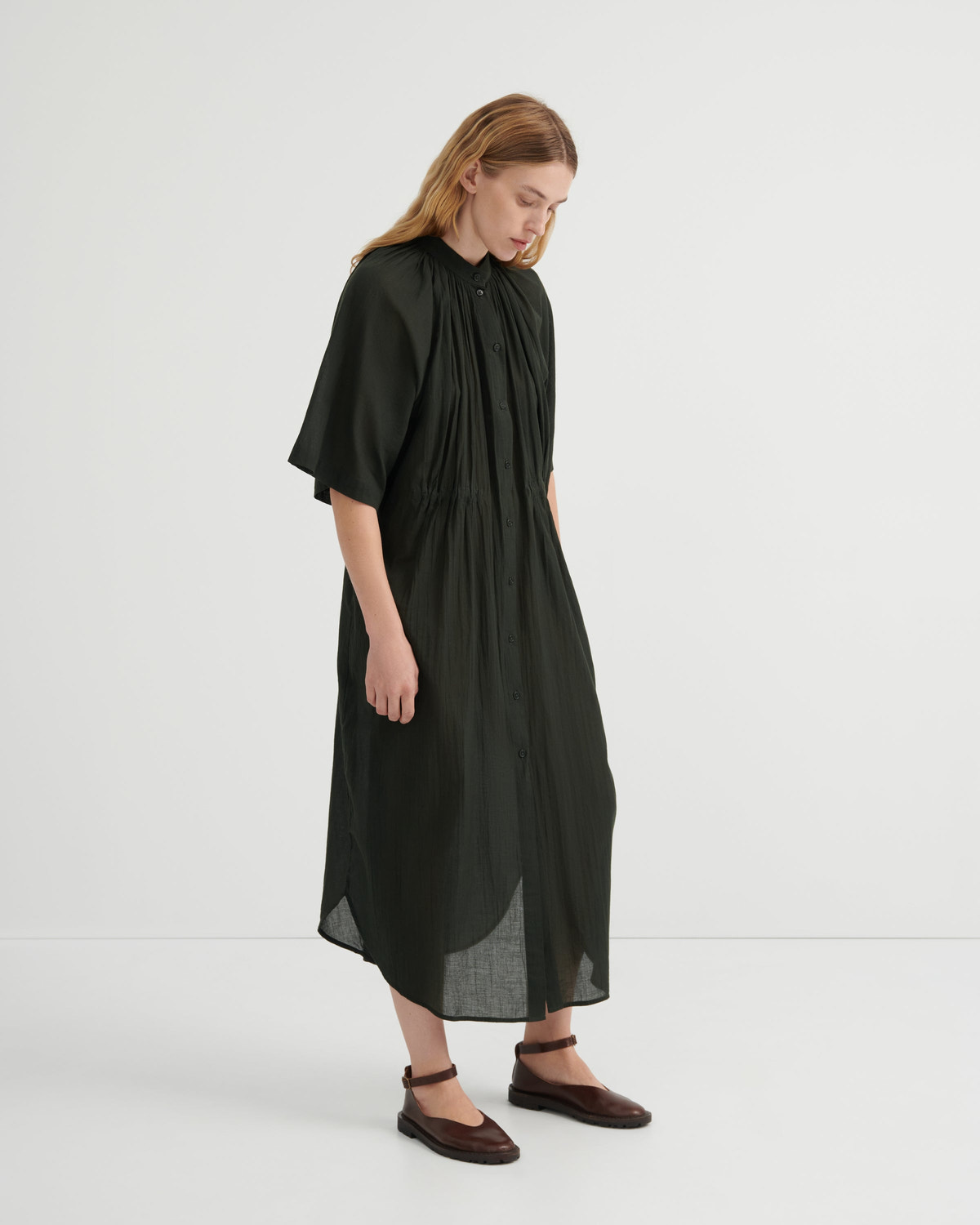 A woman modeling a loose, olive green midi dress with short flutter sleeves, a gathered high neckline, and a draped silhouette, paired with dark brown flat shoes against a plain white studio background.