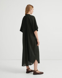 A woman stands in profile wearing a loose, flowing dark olive green dress with short wide sleeves and a draped silhouette, paired with brown flat shoes, against a clean light gray studio background.