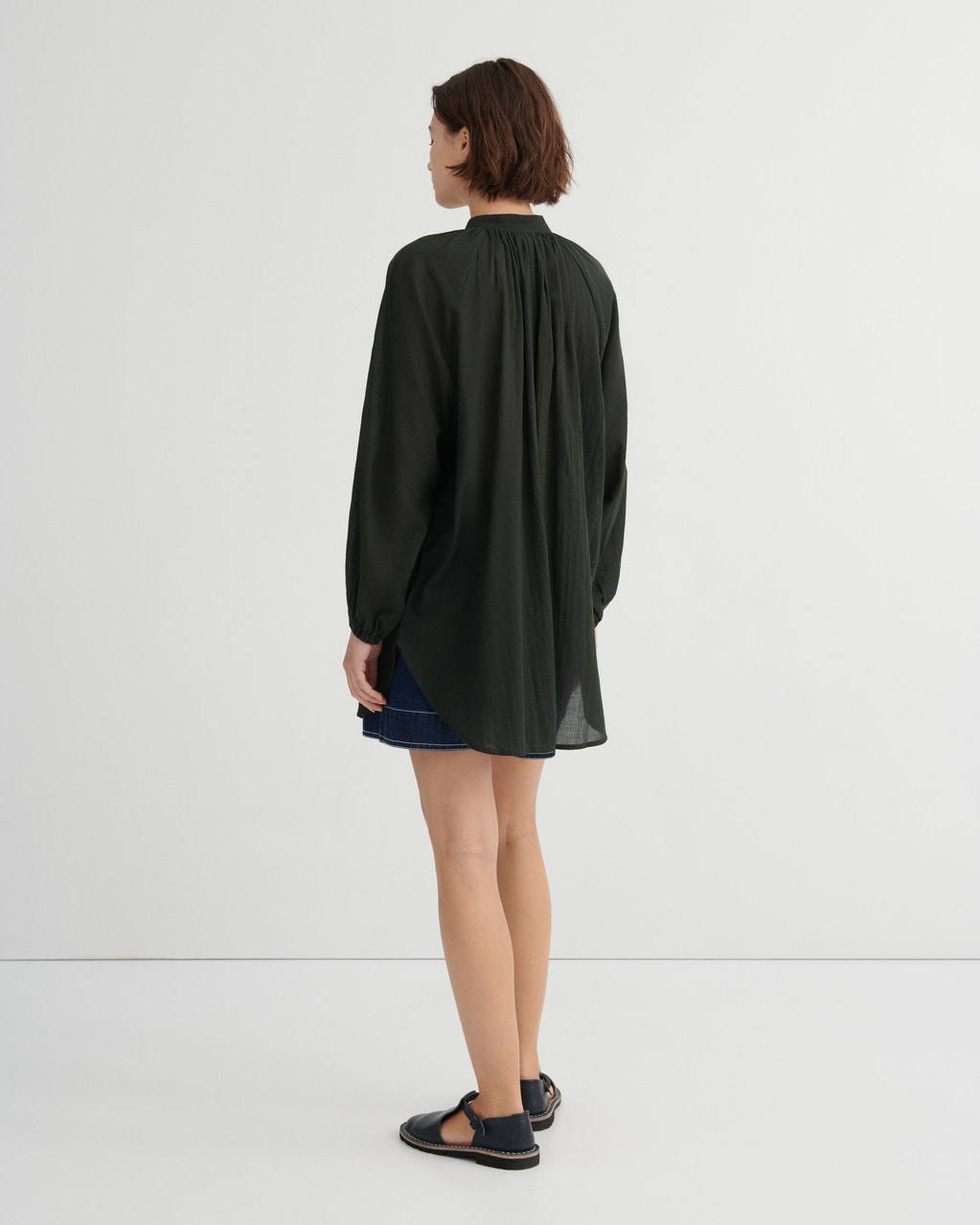 A woman shown from the back wearing a dark olive green long-sleeve blouse with a gathered neckline, paired with a dark denim skirt and black sandals, against a plain white studio background.