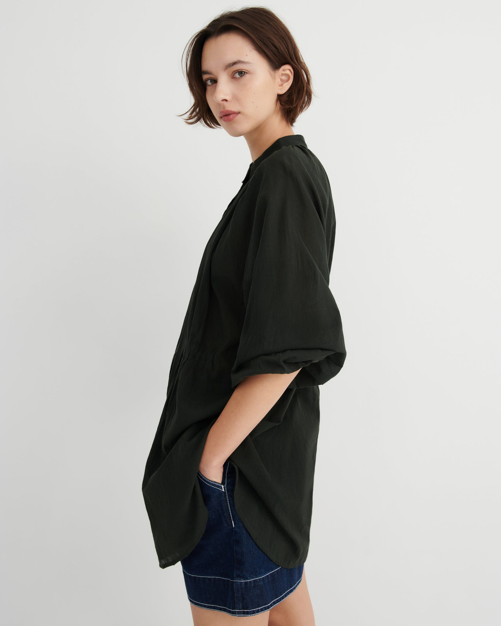 A woman wearing an oversized black short-sleeve button-up shirt with rolled cuffs and a dark denim skirt, photographed against a plain light gray studio background.