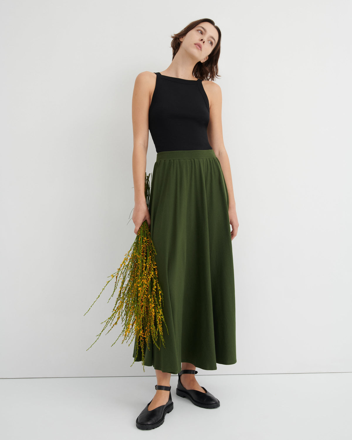 Woman wearing a black top and green skirt holding flowers against a white background