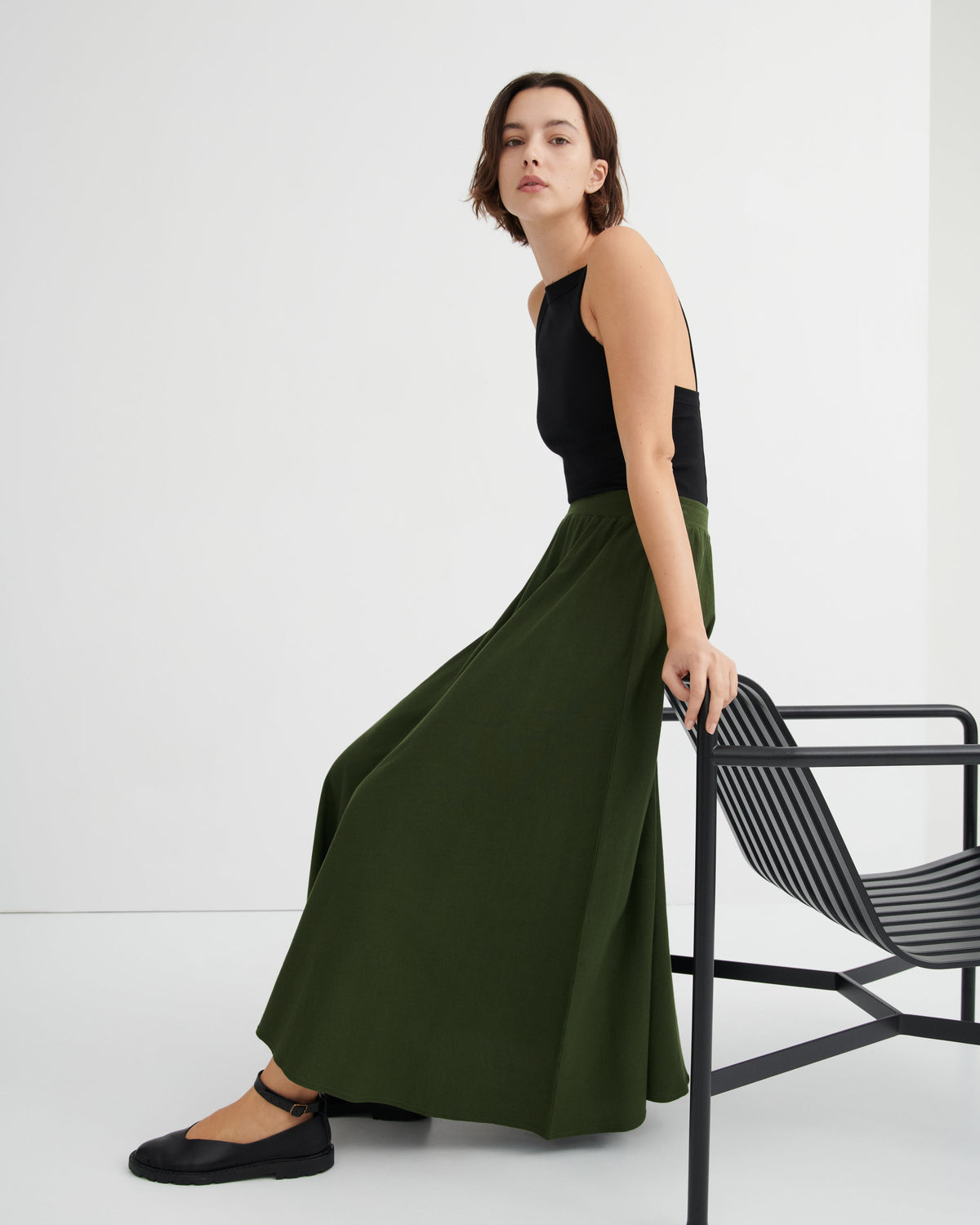 A woman models a dark olive green wide-leg skirt paired with a black sleeveless top and black flat shoes, leaning against a black metal chair in a minimalist studio setting.