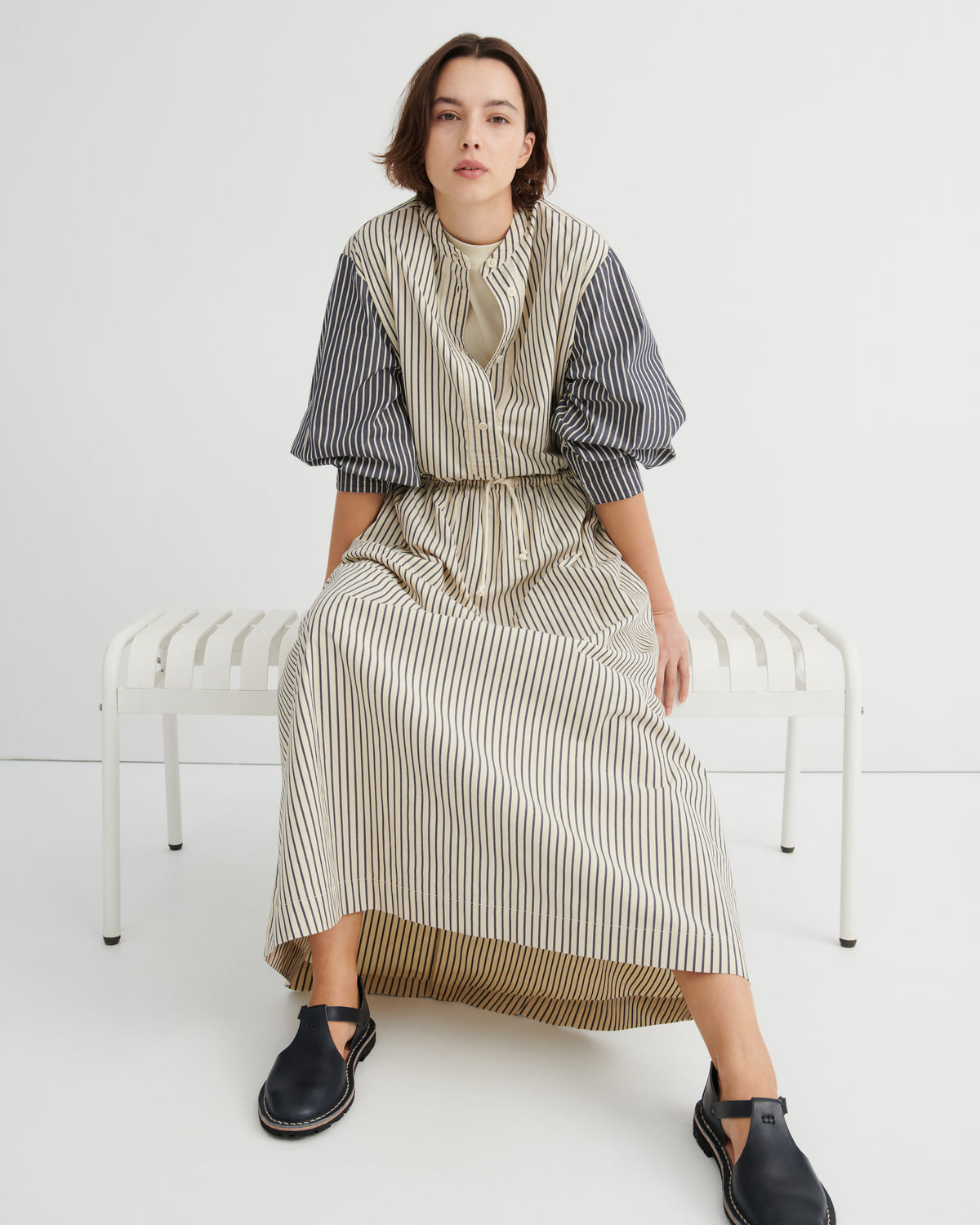 A woman sits on a white bench wearing a long-sleeved midi dress with a beige and white striped body and navy blue and white striped sleeves, paired with black Mary Jane shoes, against a plain white studio background.