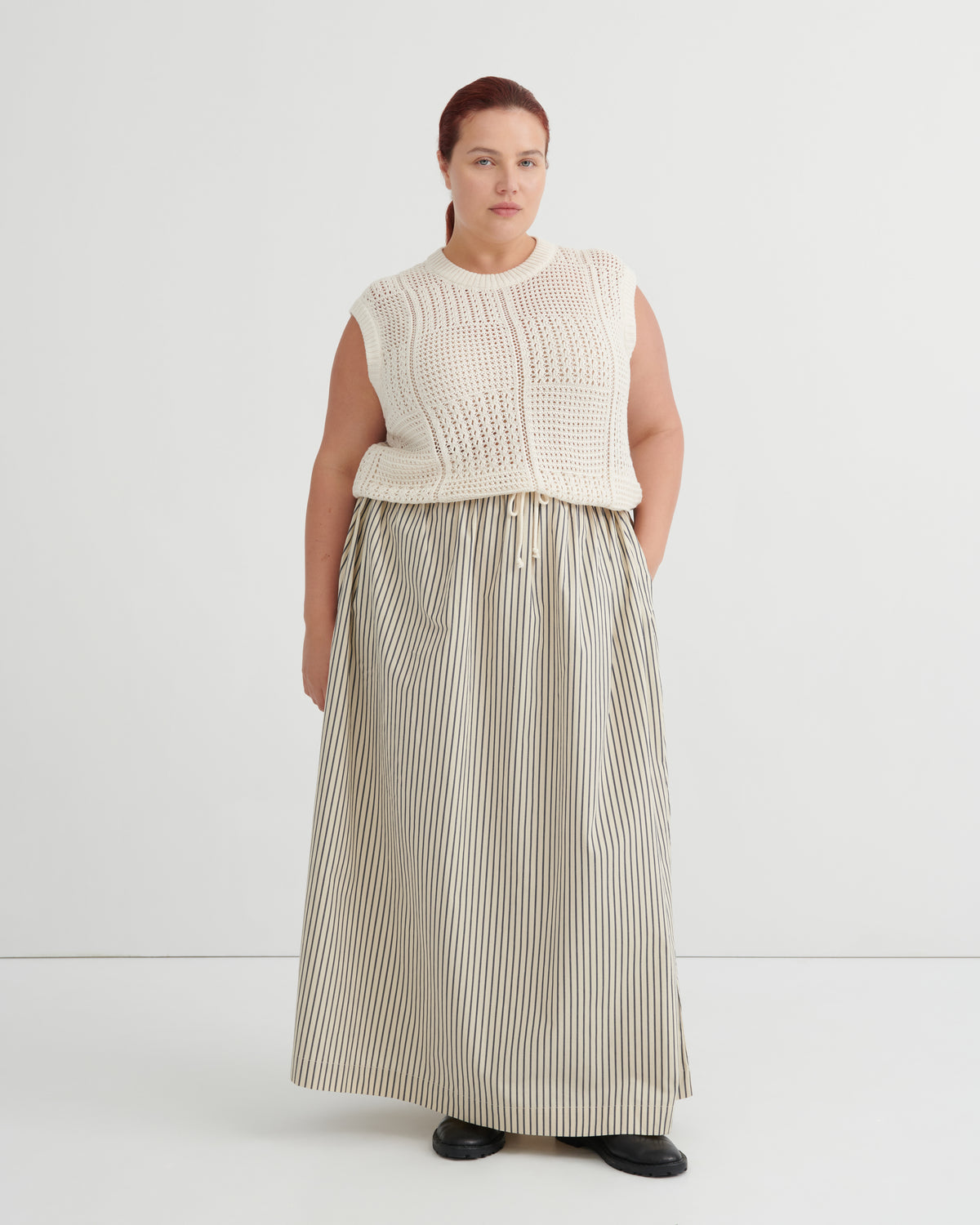 A woman modeling a cream sleeveless knit top paired with a beige and white vertically striped maxi skirt, standing against a plain white studio background.