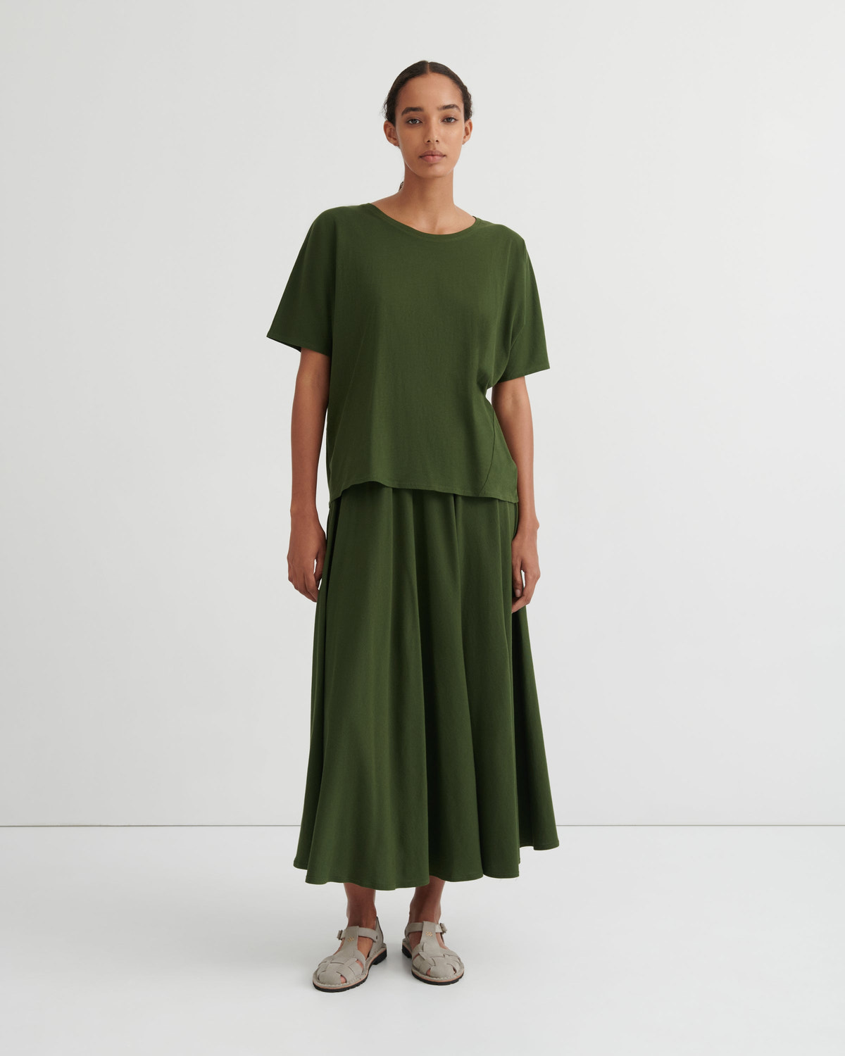 A model stands against a plain white studio background wearing a matching olive green two-piece outfit consisting of a short-sleeve top and a long pleated skirt, paired with light-colored flat sandals.