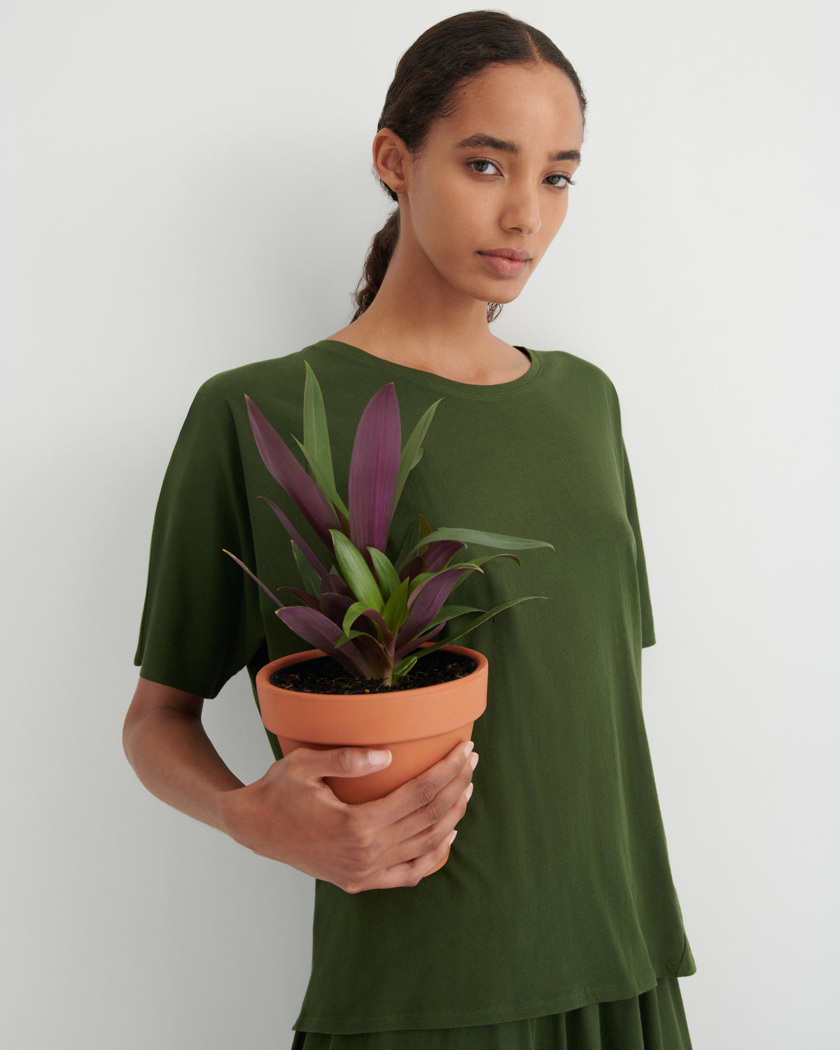 A woman wearing an olive green short-sleeve top holds a potted plant with purple and green leaves in a terracotta pot, set against a plain light gray background.