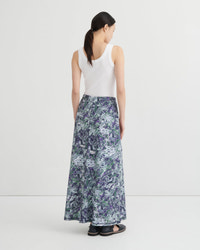 A woman shown from the back wearing a sleeveless white top and a long marbled skirt with a blue, purple, and green pattern, standing against a plain light gray studio background.