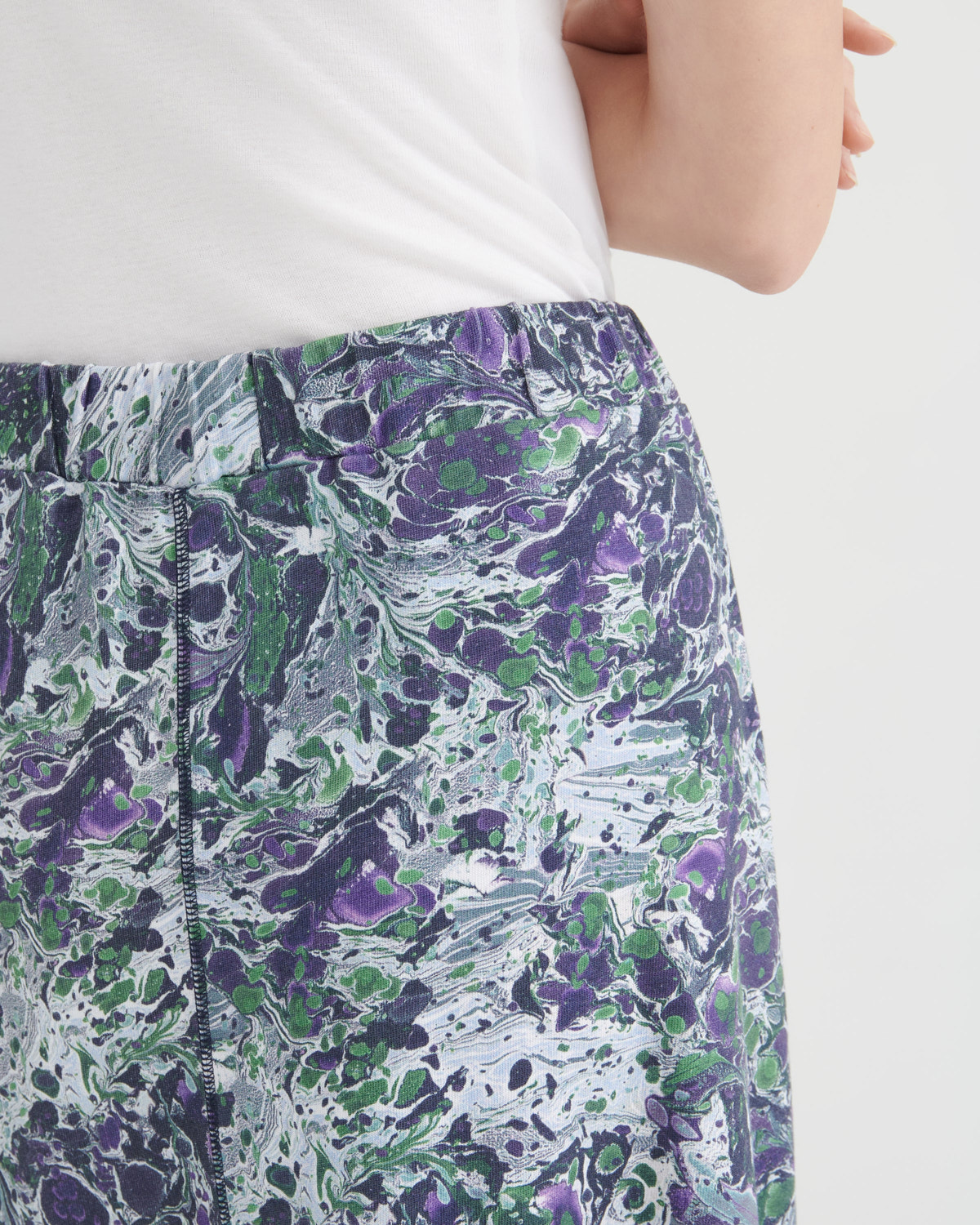 Close-up of a person wearing patterned skirt with an abstract marbled print in purple, green, black, and white, paired with a white top against a light gray background.