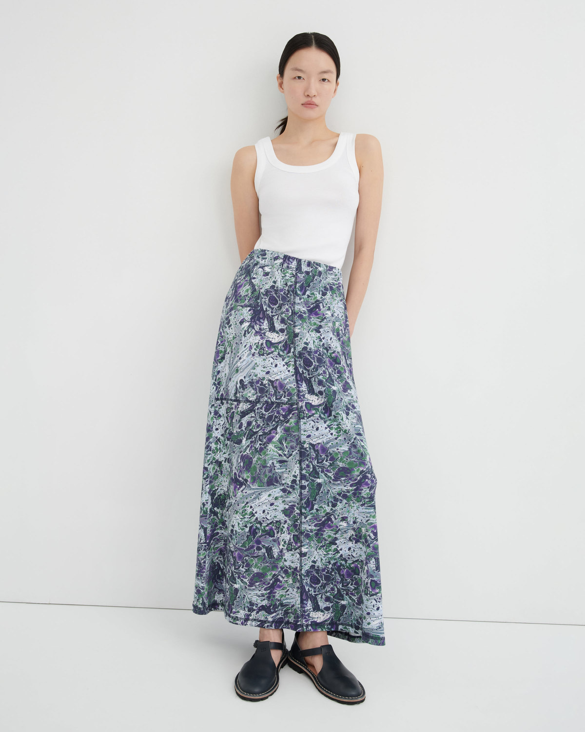 A model stands against a plain white studio background wearing a white ribbed tank top, a long high-waisted maxi skirt with a blue, purple, and white abstract marbled print, and black flat shoes.