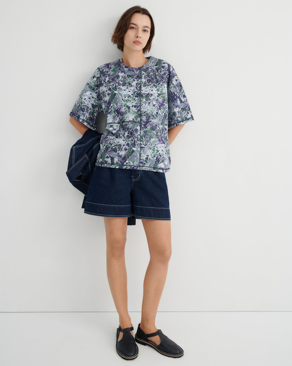 A model stands against a plain light gray studio wall wearing a short-sleeve button-front shirt with a blue, green, and white marbled print, dark denim shorts, and black slip-on shoes. A dark blue jacket is draped over one shoulder.