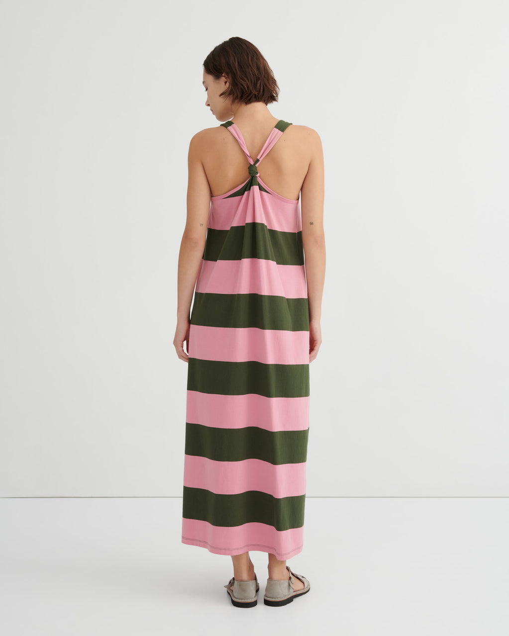 A woman viewed from the back wearing a sleeveless maxi dress with wide horizontal pink and dark green stripes and a crossover halter-style back, against a clean white studio background.