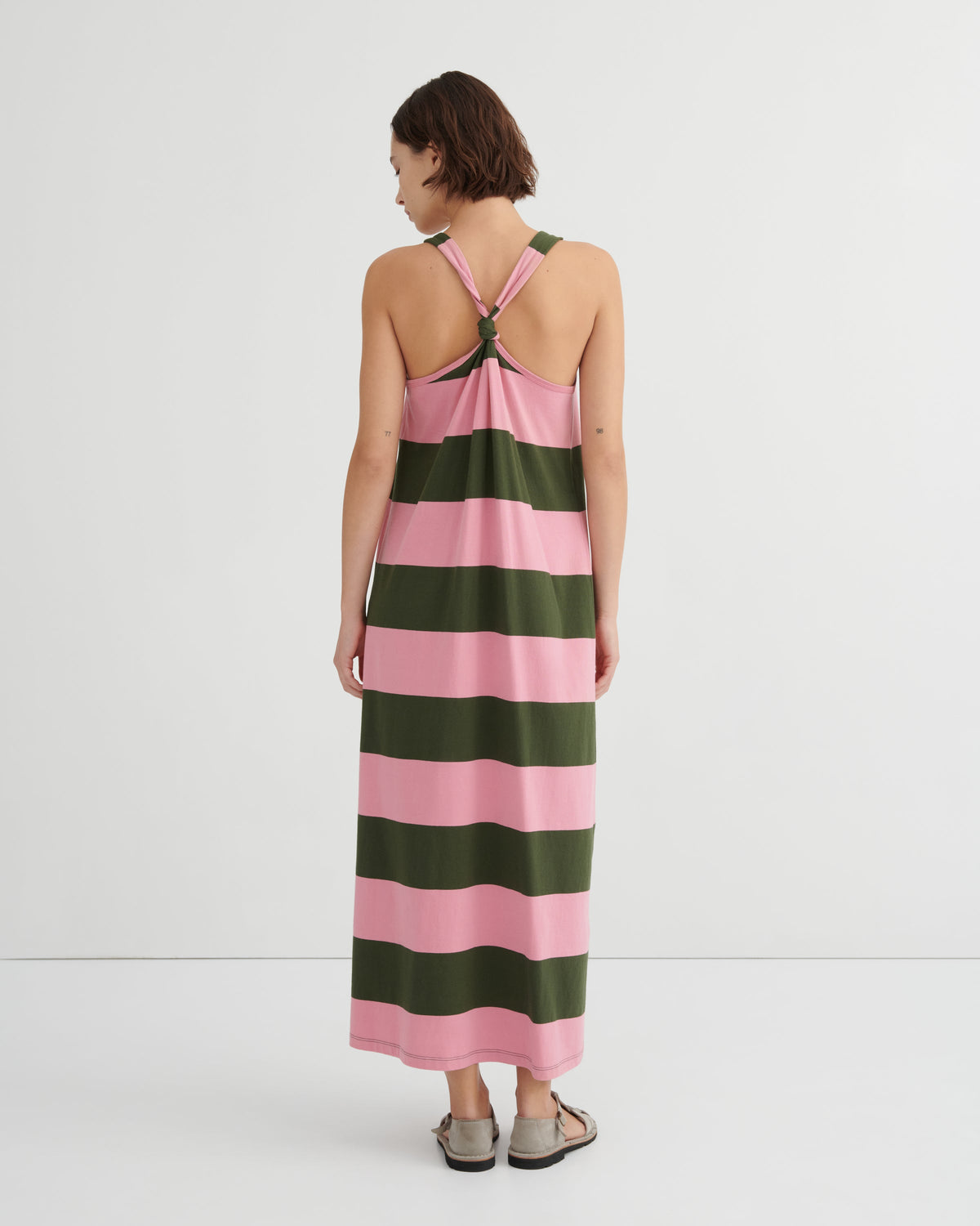 A woman viewed from the back wearing a sleeveless maxi dress with wide horizontal pink and dark green stripes and a crossover halter-style back, against a clean white studio background.