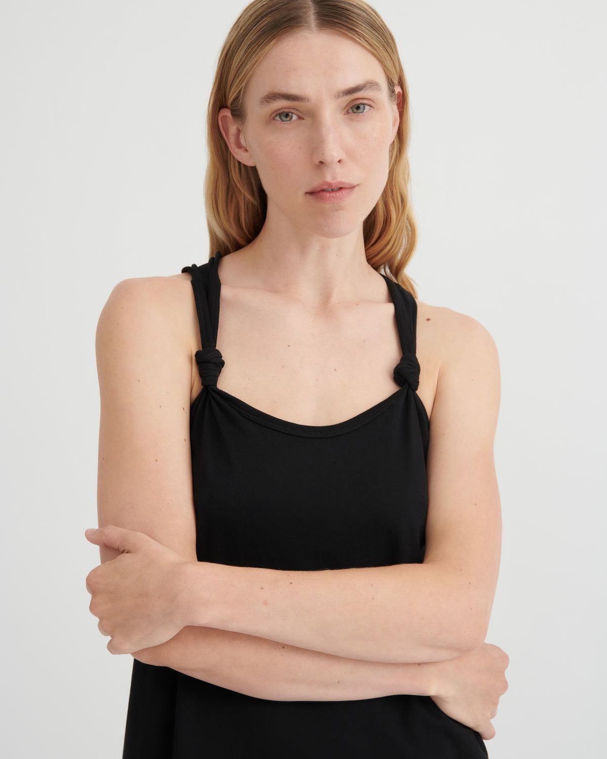 A woman modeling a black camisole tank top with knotted shoulder straps, photographed against a clean light gray studio background.