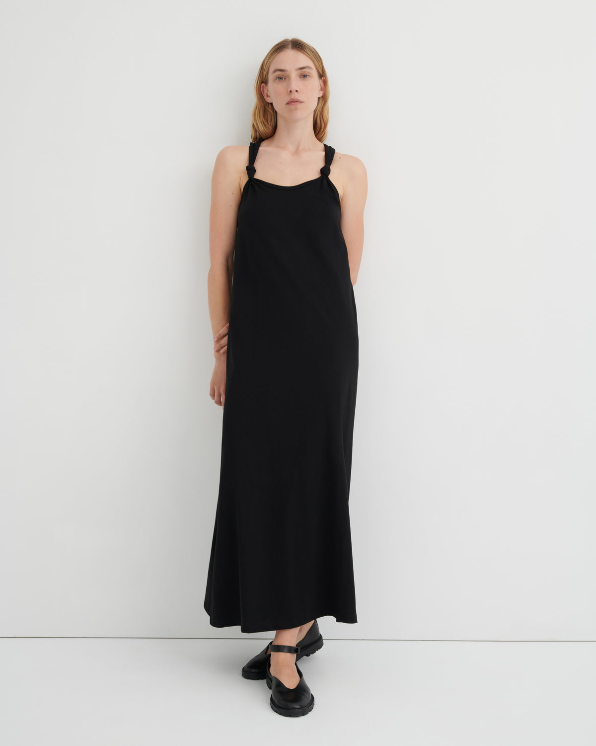 A woman modeling a black sleeveless halter-neck maxi dress with a subtle sheen, paired with black Mary Jane shoes, against a clean white studio background.
