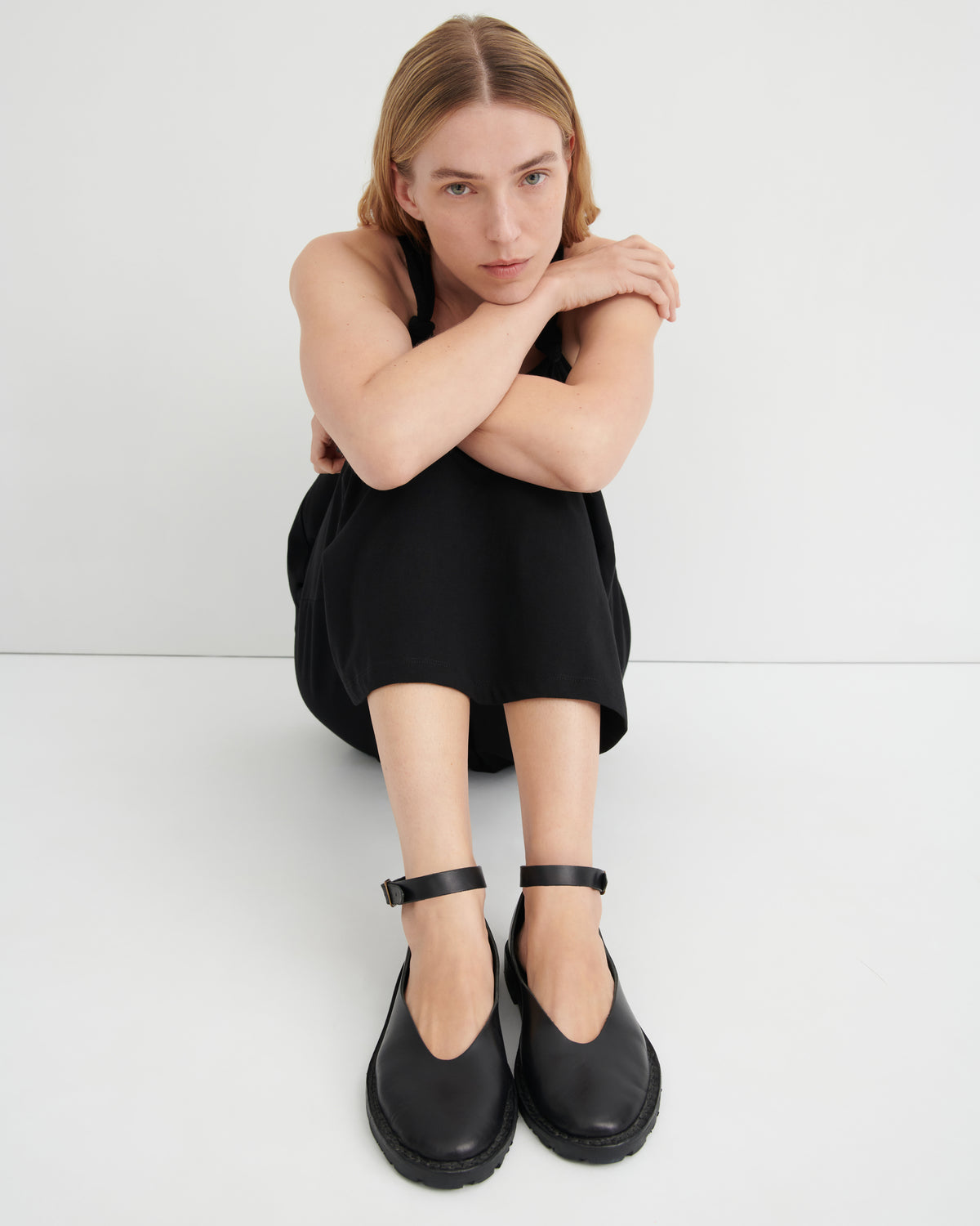 A model sits cross-legged on a light gray studio floor wearing a black sleeveless dress and black Mary Jane-style shoes with ankle straps.