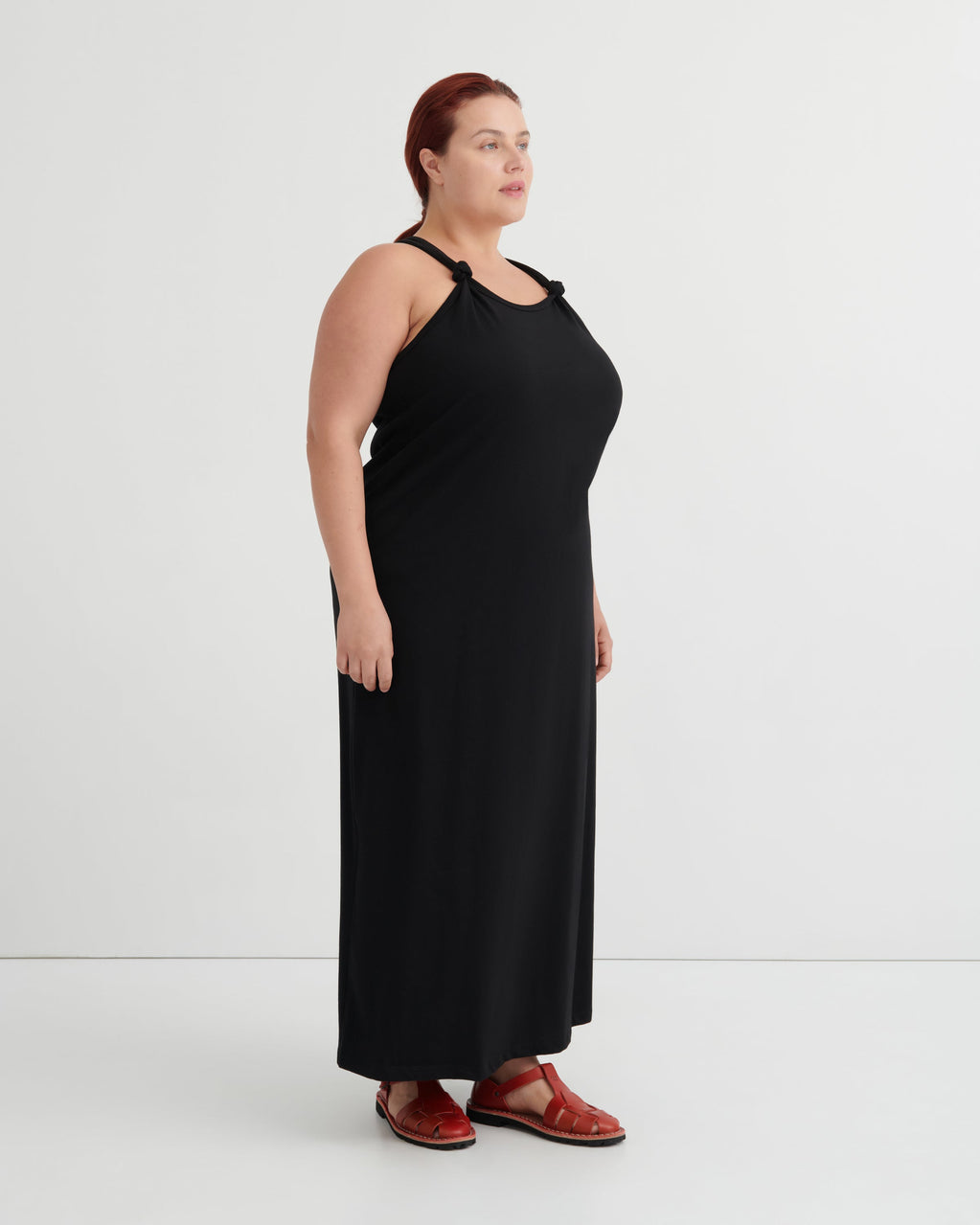 A woman modeling a black halter-neck maxi dress with shoulder cutouts, paired with brown leather sandals, standing against a plain white studio background.