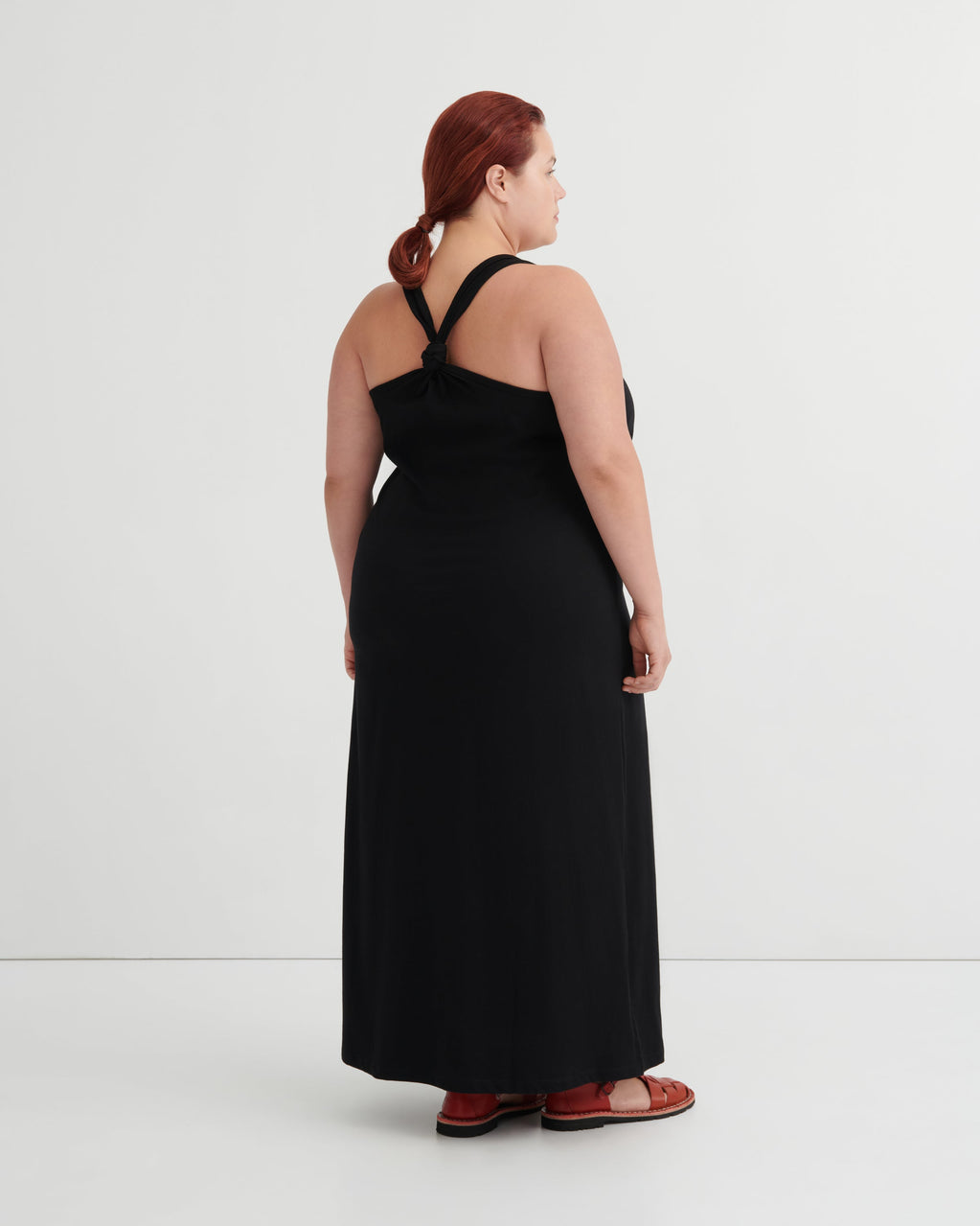 A woman with red hair tied back is shown from the back wearing a black halter-neck maxi dress and brown sandals against a plain white studio background.