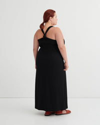 A woman with red hair tied back is shown from the back wearing a black halter-neck maxi dress and brown sandals against a plain white studio background.