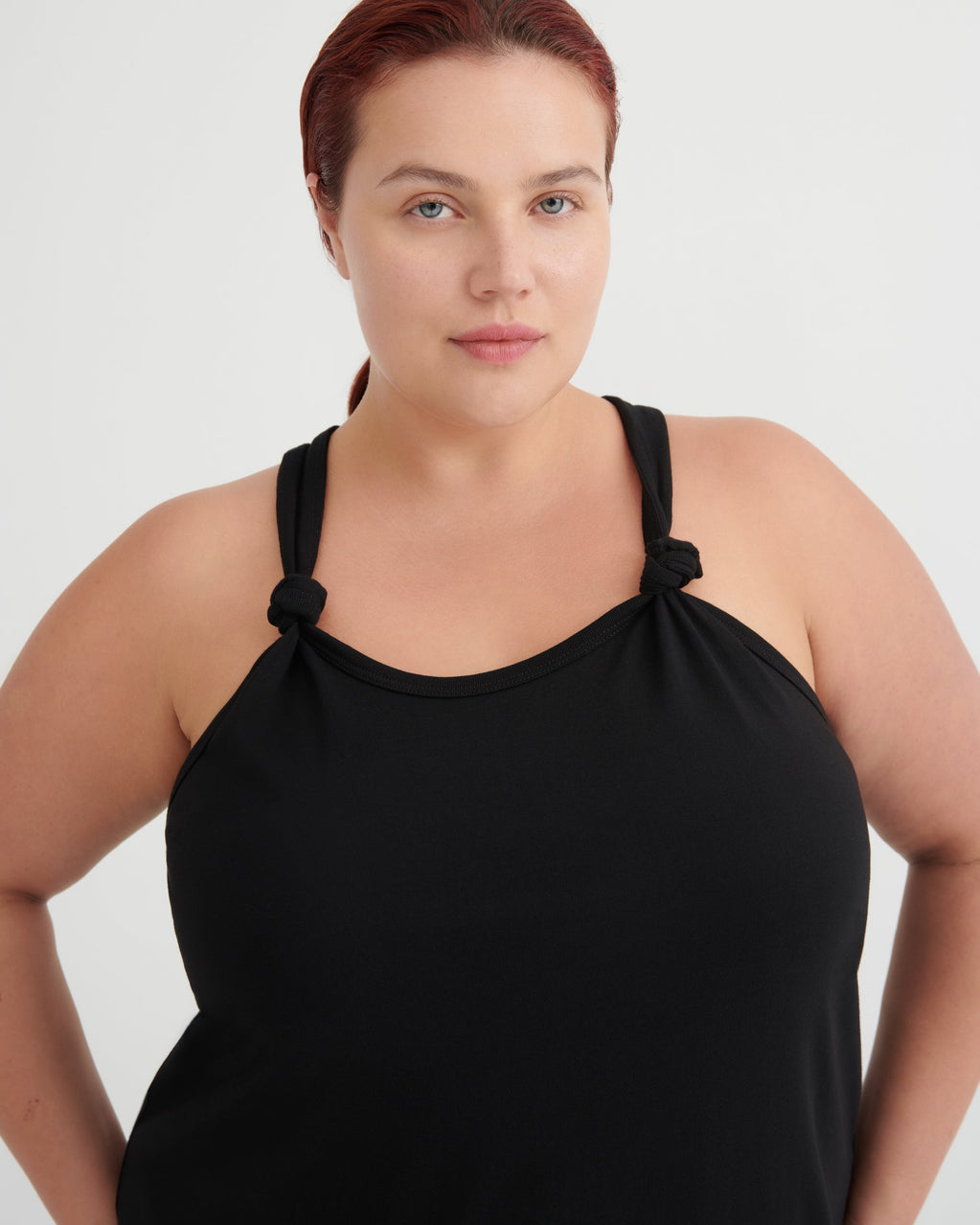 A plus-size model with red hair tied back, wearing a black halter-style tank top with knotted shoulder details, photographed against a light gray studio background.