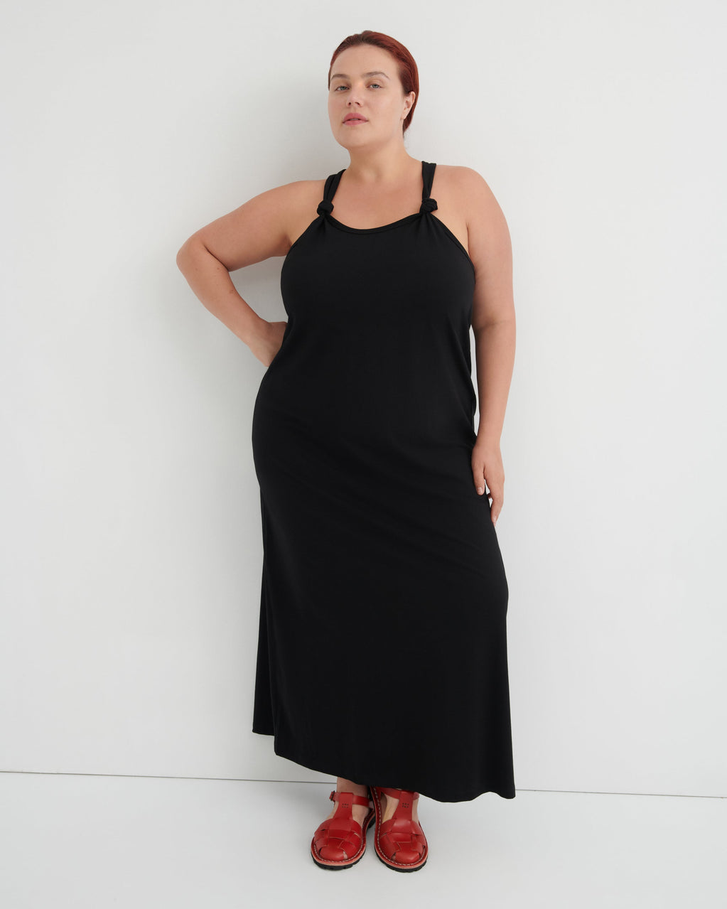 A woman modeling a black halter-neck maxi dress with shoulder cutouts, paired with brown leather sandals, standing against a plain white studio background.