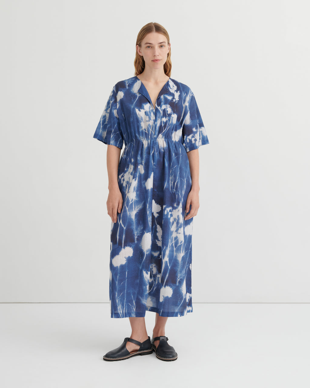 A woman stands against a plain light gray studio background wearing a loose, short-sleeve midi dress with a blue and white tie-dye or shibori-style abstract pattern. The dress has a V-neck and gathered waist.