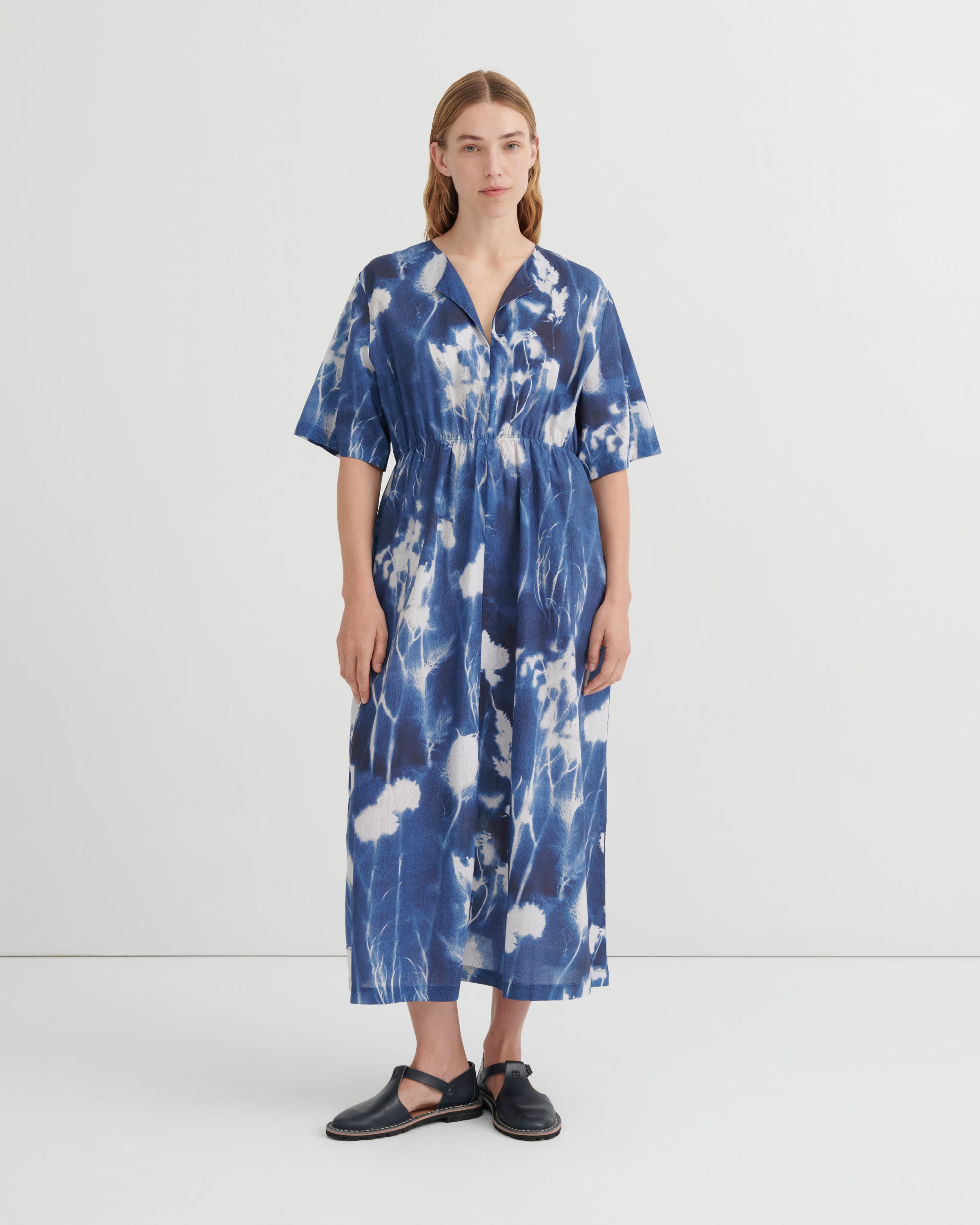 A woman stands against a plain light gray studio background wearing a loose, short-sleeve midi dress with a blue and white tie-dye or shibori-style abstract pattern. The dress has a V-neck and gathered waist.