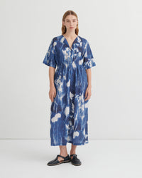 A woman stands against a plain light gray studio background wearing a loose, short-sleeve midi dress with a blue and white tie-dye or shibori-style abstract pattern. The dress has a V-neck and gathered waist.