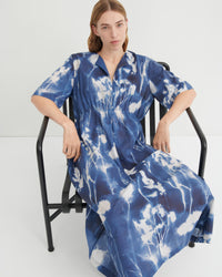 A woman wearing a blue and white tie-dye kaftan dress sits on a black metal chair against a plain white studio background.