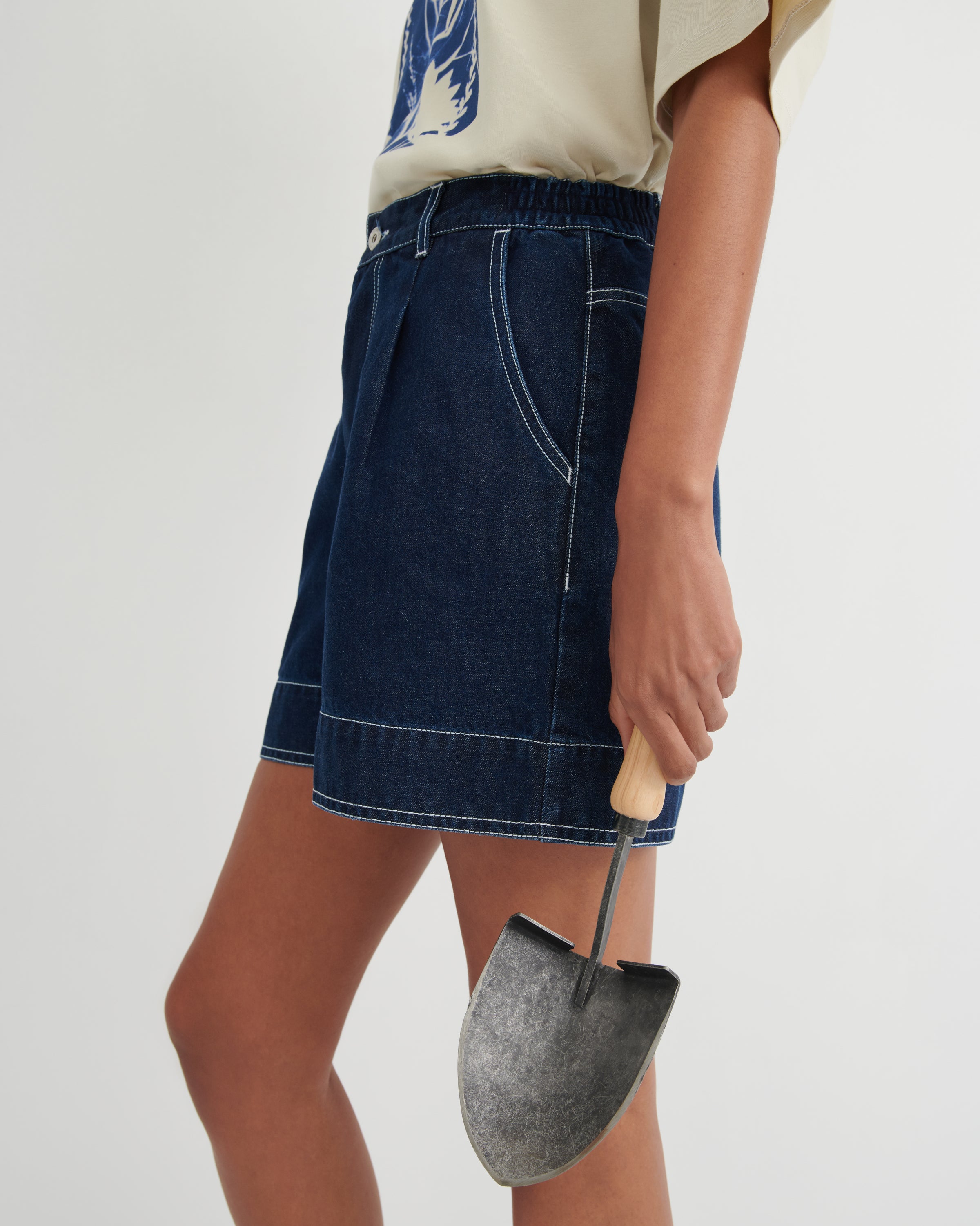 A model wearing a dark blue denim shorts with contrast white stitching, holding a small metal trowel, against a plain light gray studio background.