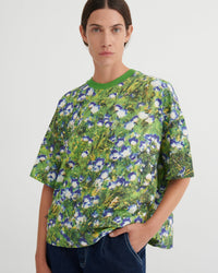 A model wearing a green short-sleeve crewneck t-shirt with a dense blue, white, and yellow floral print, paired with dark blue jeans, against a plain light gray background.