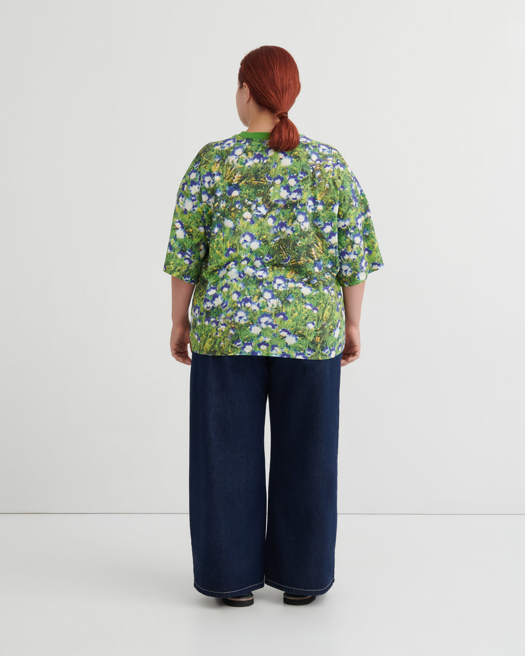 A person viewed from the back wearing a green short-sleeve top with a dense blue and white floral print, paired with dark wide-leg pants, against a plain light gray studio background.