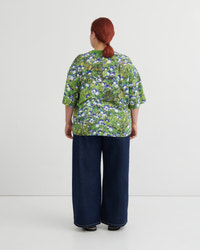 A person viewed from the back wearing a green short-sleeve top with a dense blue and white floral print, paired with dark wide-leg pants, against a plain light gray studio background.