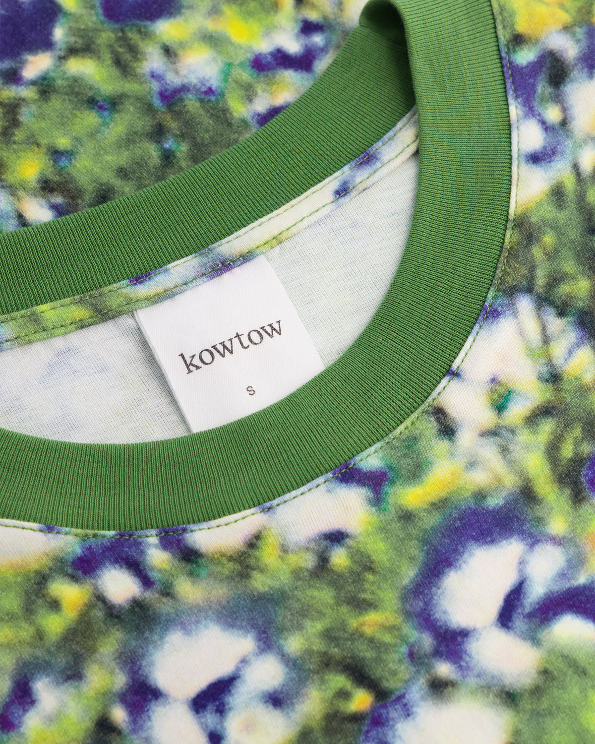 Close-up of a t-shirt neck showing a green ribbed collar and a white brand label that reads 'kowtow'. The shirt features a vibrant green, purple, and yellow tie-dye pattern.