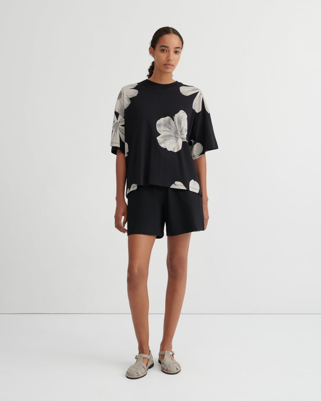 A model stands against a plain white studio background wearing a black short-sleeve top with large white floral prints and matching black shorts.
