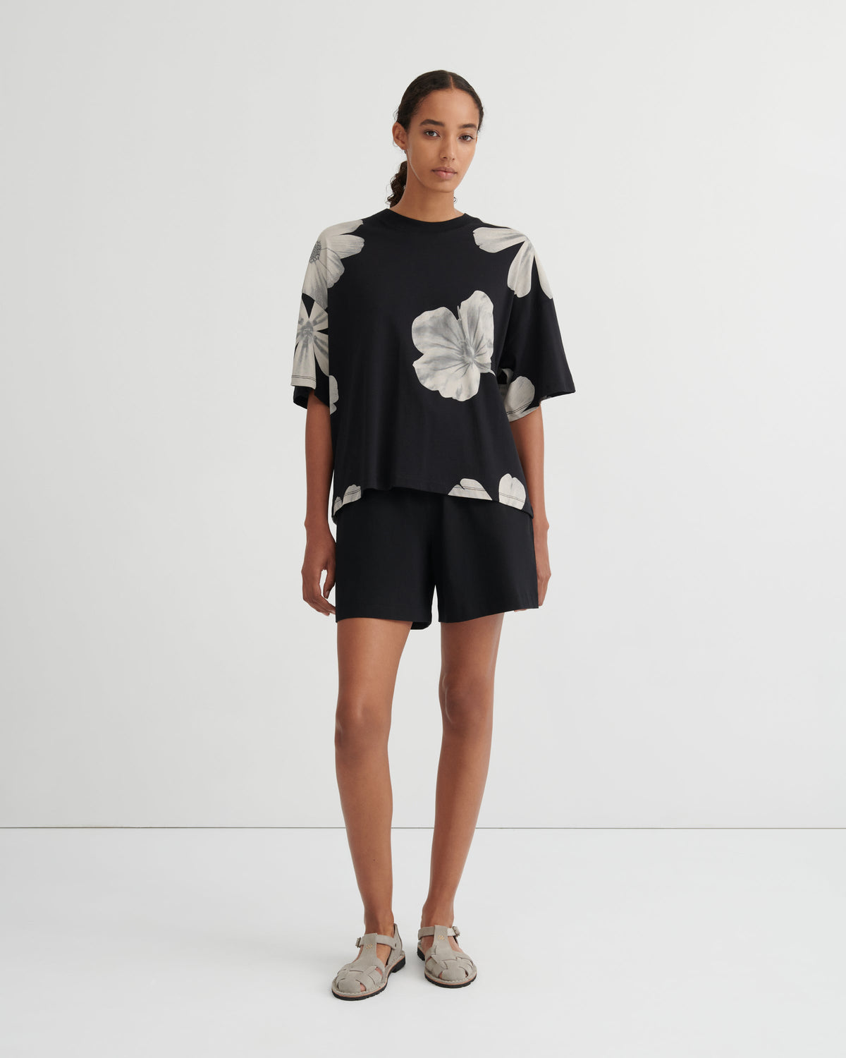 A model stands against a plain white studio background wearing a black short-sleeve top with large white floral prints and matching black shorts.