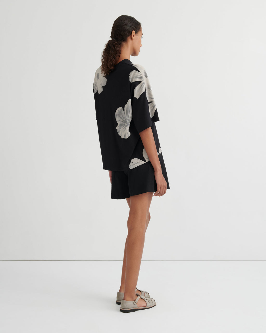 A model stands against a plain white studio background wearing a black short-sleeve top with large white floral prints and matching black shorts.