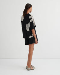 A model stands against a plain white studio background wearing a black short-sleeve top with large white floral prints and matching black shorts.