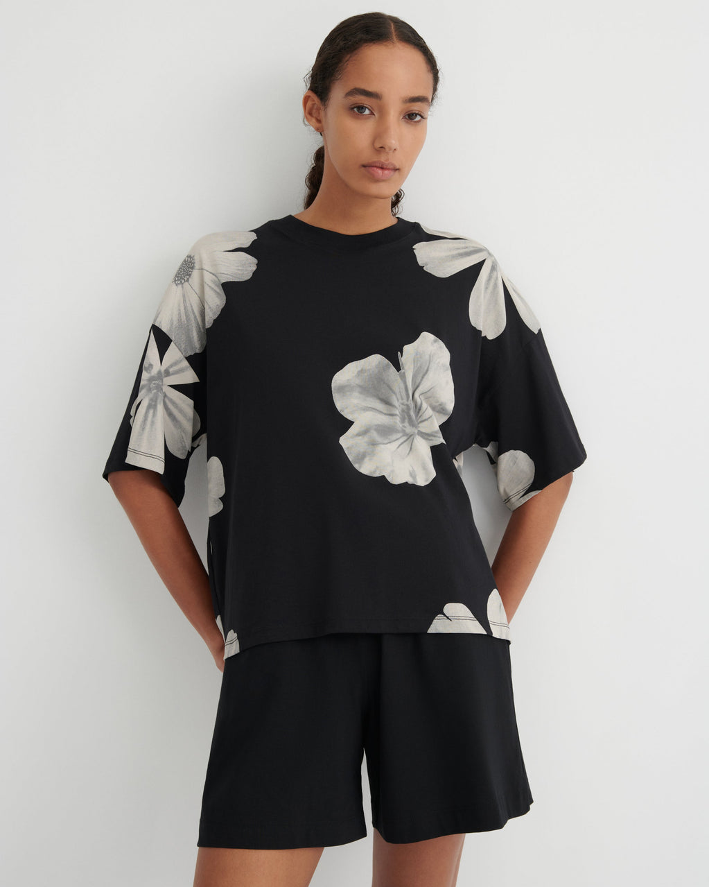 A woman modeling a black short-sleeve top with large white floral prints on the sleeves and chest, paired with black shorts, against a plain light gray background.