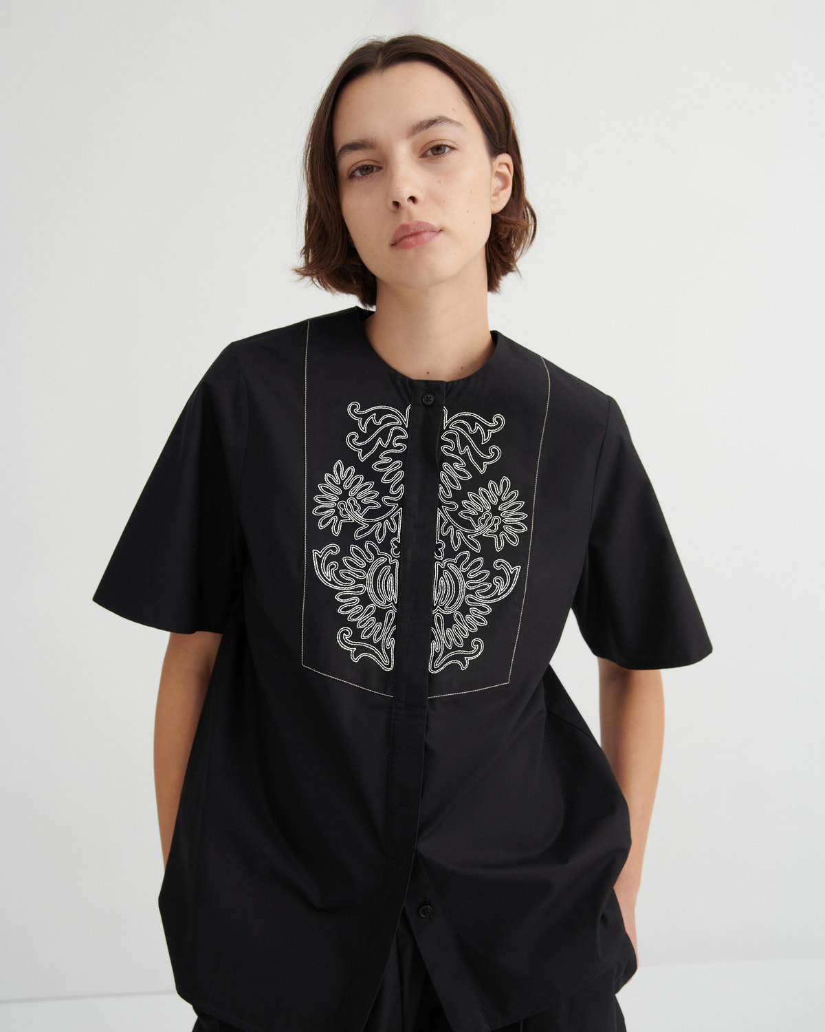 A model wearing a black short-sleeve button-up shirt with white embroidered floral details on the chest, photographed against a light gray studio background.