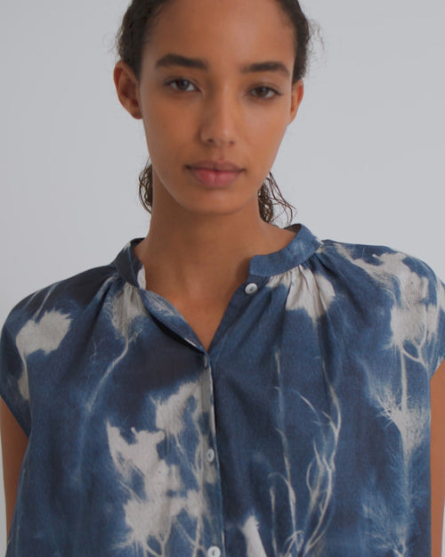 A women wears a a cyanotype print sleeveless blouse and dark blue denim shorts with brown flats.