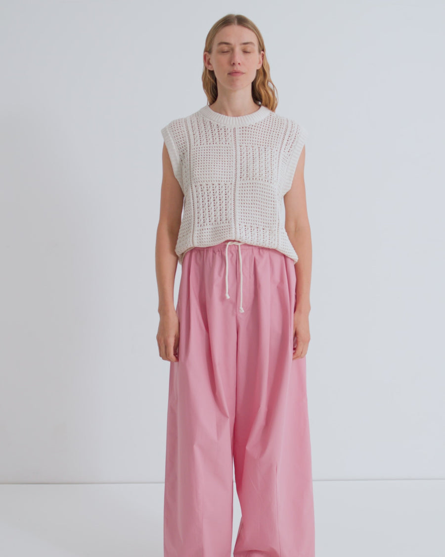 A women wearing a sleeveless crochet off white vest with pink drawcord pleated pants.