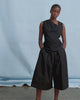 A women wearing a black asymmetric sleeveless top and black wide leg pants in front of blue backdrop.