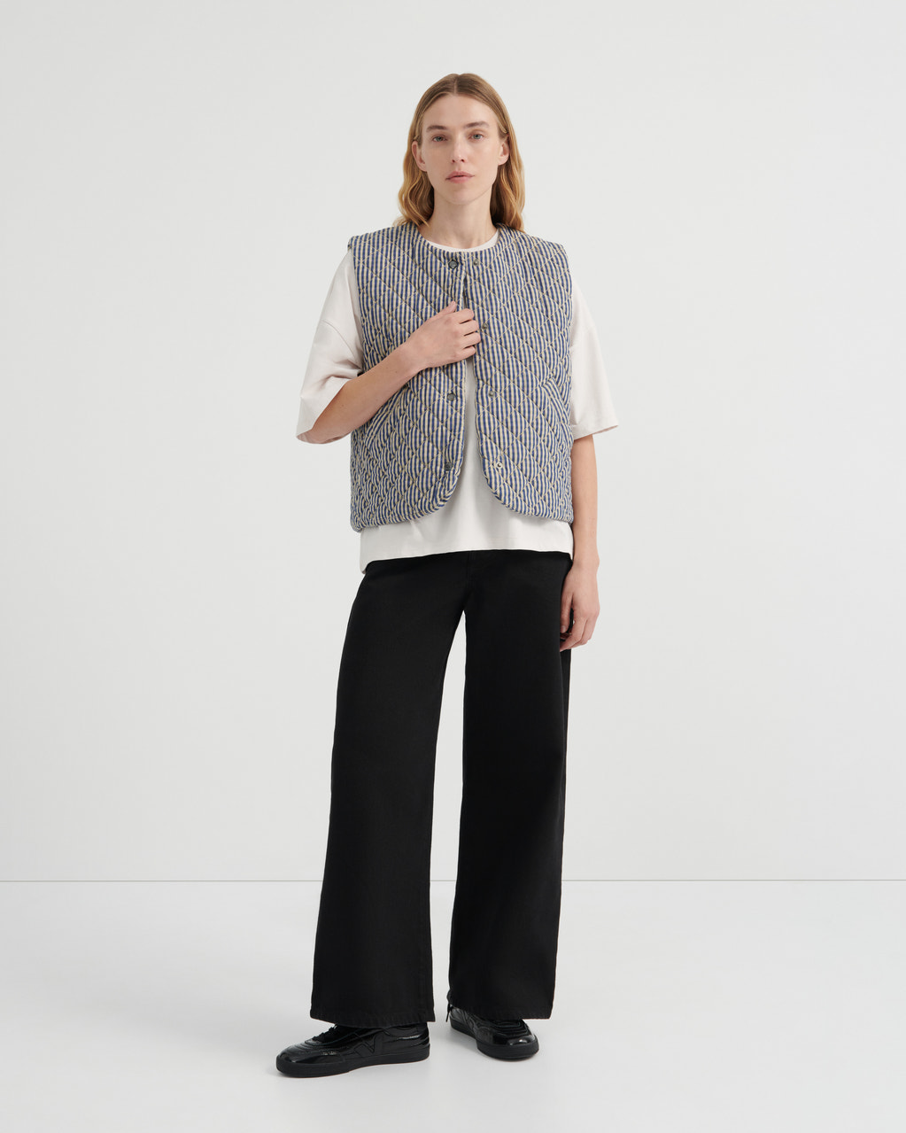 A model stands against a plain white studio background wearing a sleeveless textured gray vest over a white short-sleeve top, black wide-leg trousers, and black shoes.