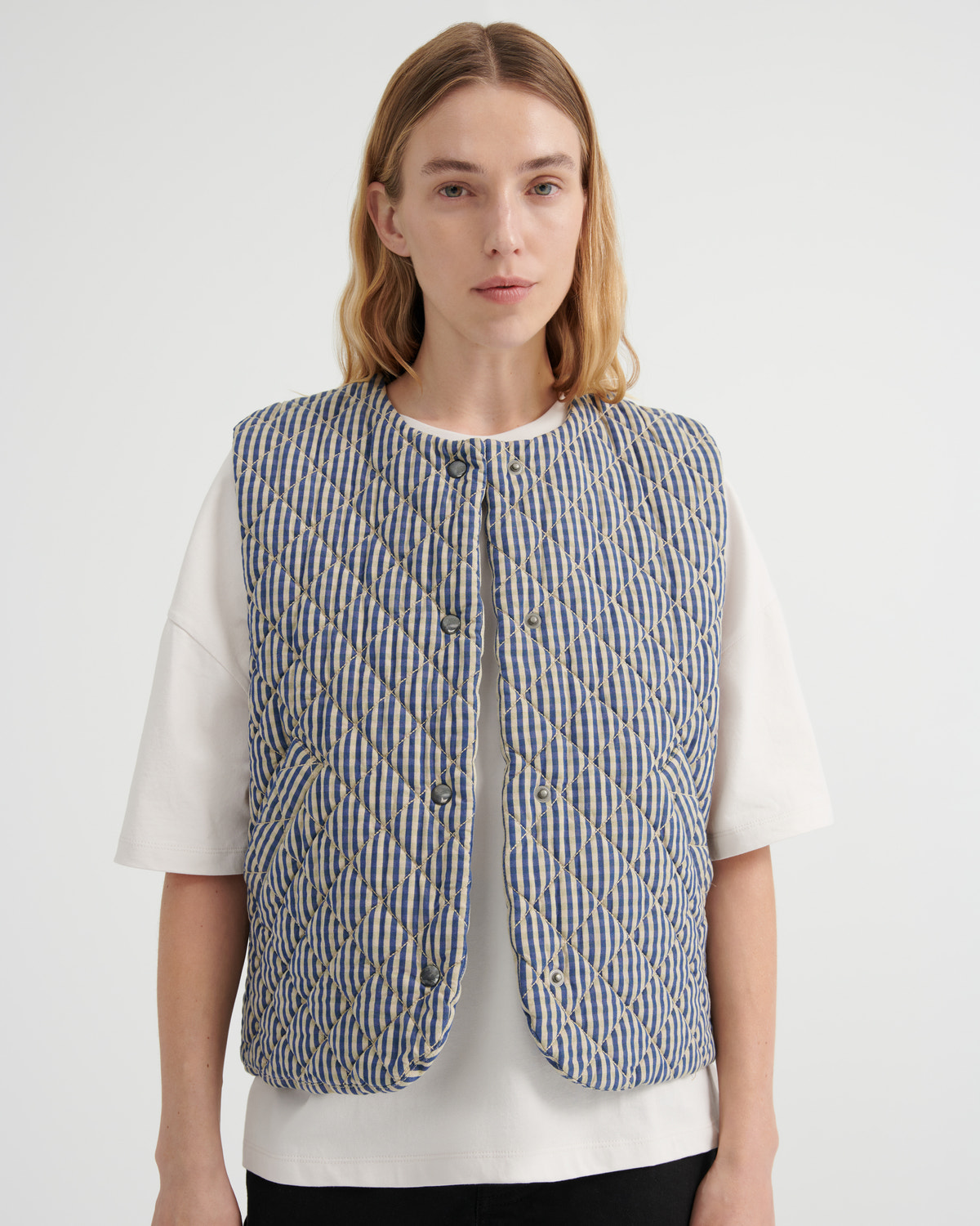A model wearing a blue and beige vertically striped quilted vest over a white t-shirt, photographed against a light gray studio background.