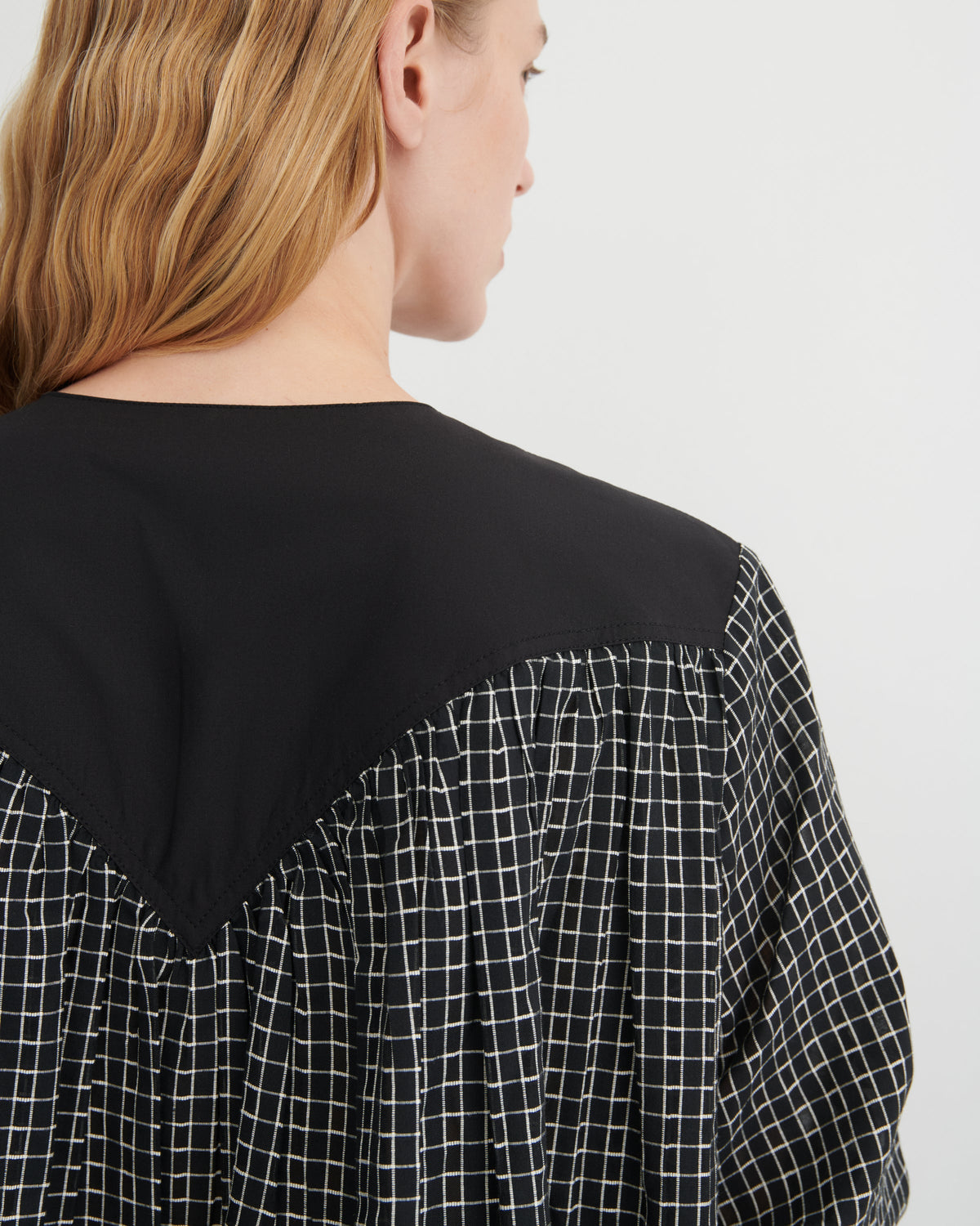 Close-up back view of a woman wearing a black top with a white windowpane grid pattern on the lower back and sleeves, against a plain light gray background.