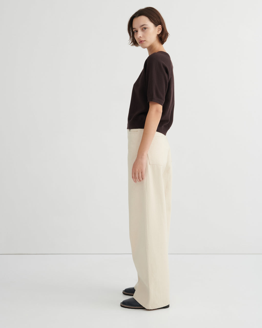 A woman stands in profile wearing a dark brown short-sleeve top, cream wide-leg trousers, and dark loafers against a clean white studio background.