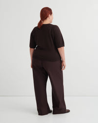 A model with red hair tied back is shown from behind wearing a dark brown short-sleeve top and matching wide-leg trousers against a light gray studio background.