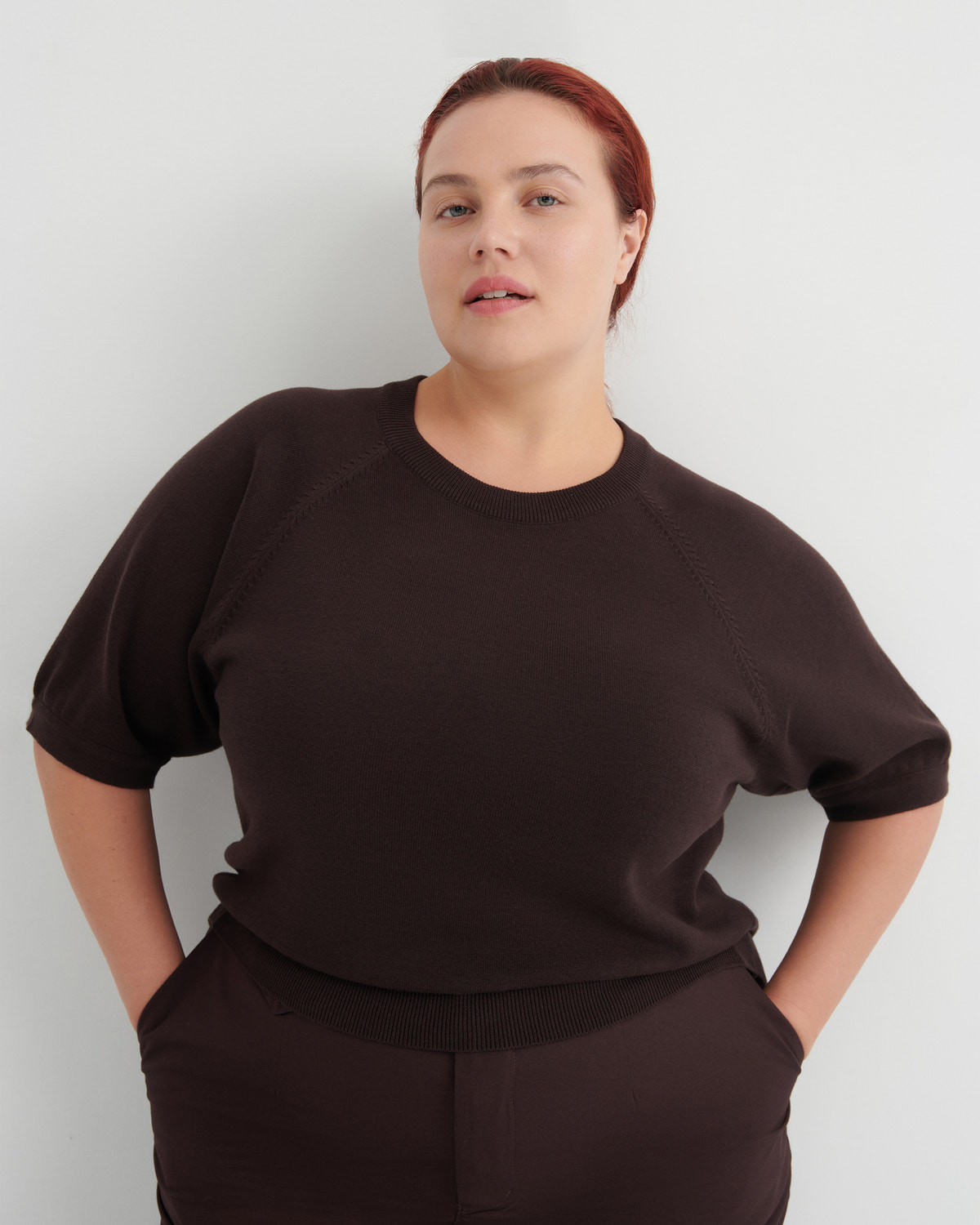 A woman with red hair pulled back wears a dark brown short-sleeve knit top and matching dark brown bottoms, standing against a light gray studio background with her hands in her pockets.
