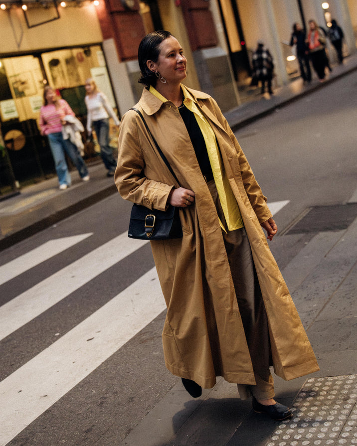 A woman walking on a city crosswalk wearing a long tan trench coat over a black and yellow top, with brown trousers and a black shoulder bag.
